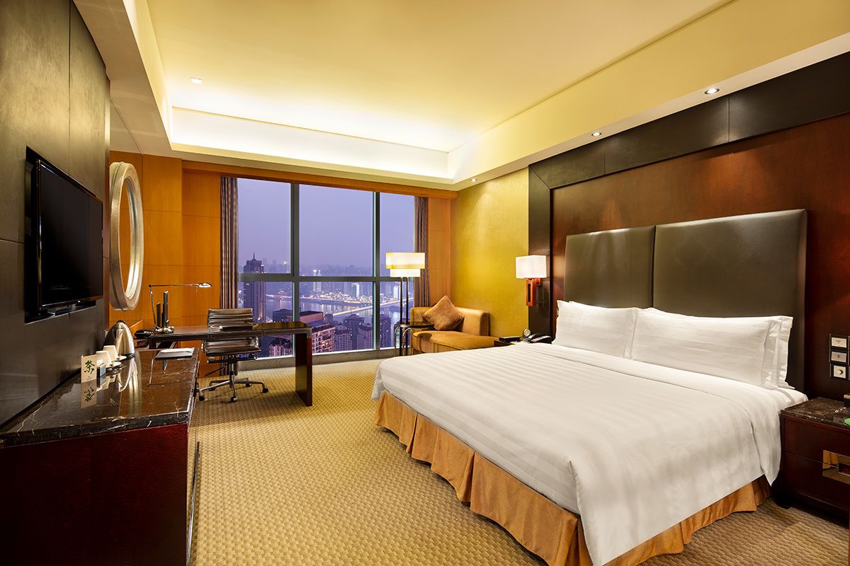 A hotel room with a large bed, a desk, a TV, and a view of a cityscape through large windows.