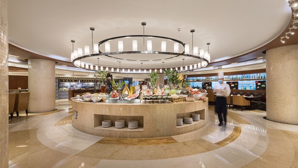 A modern hotel lobby with a central buffet area, a bar, and seating arrangements.