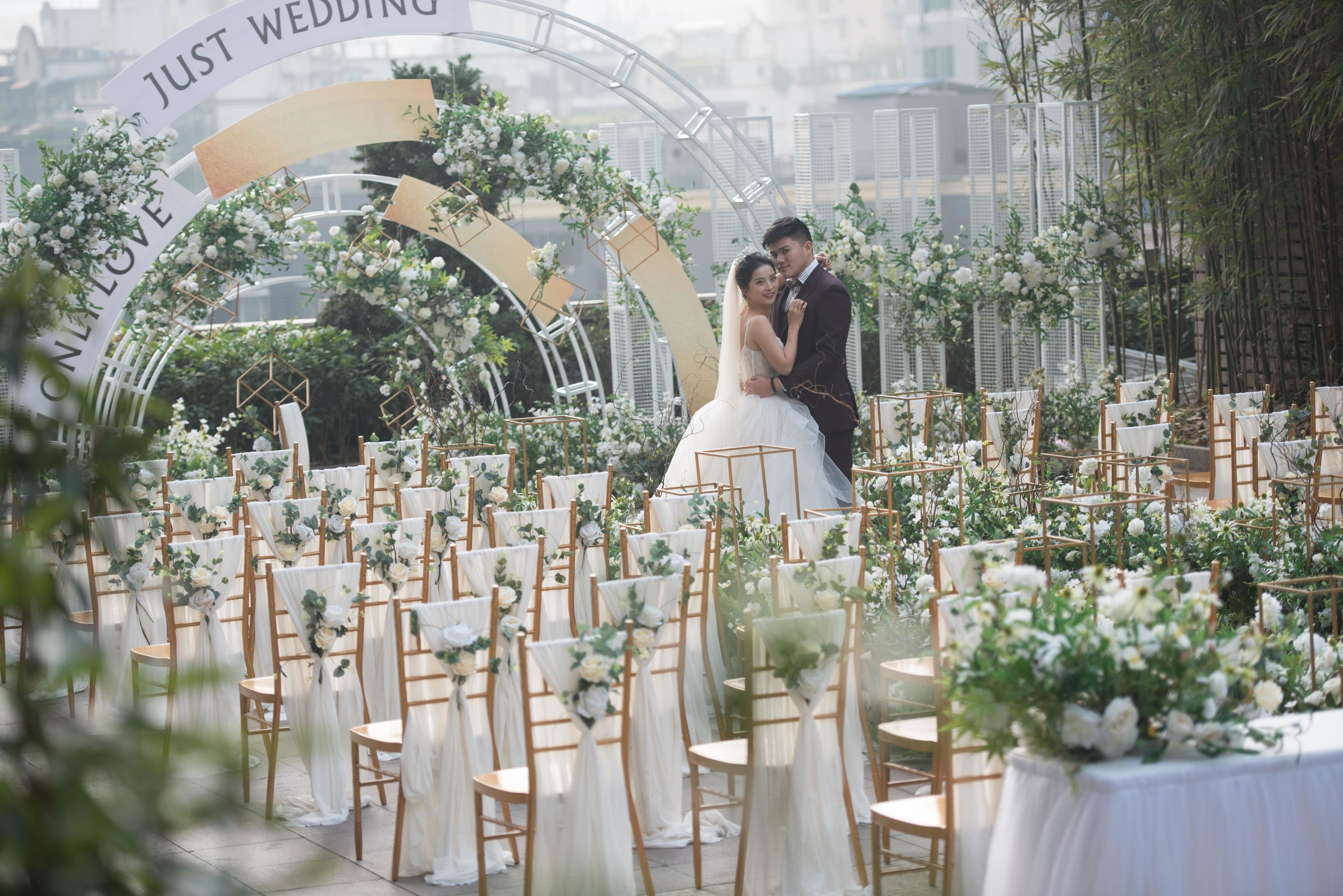 A wedding ceremony with a couple kissing under an arch with "JUST WEDDING" text. Rows of chairs with white drapes and floral arrangements are visible.