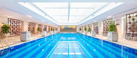 Ein großer, überdachter Swimmingpool mit hellblauen Kacheln, umgeben von Liegestühlen und Pflanzen in Töpfen.