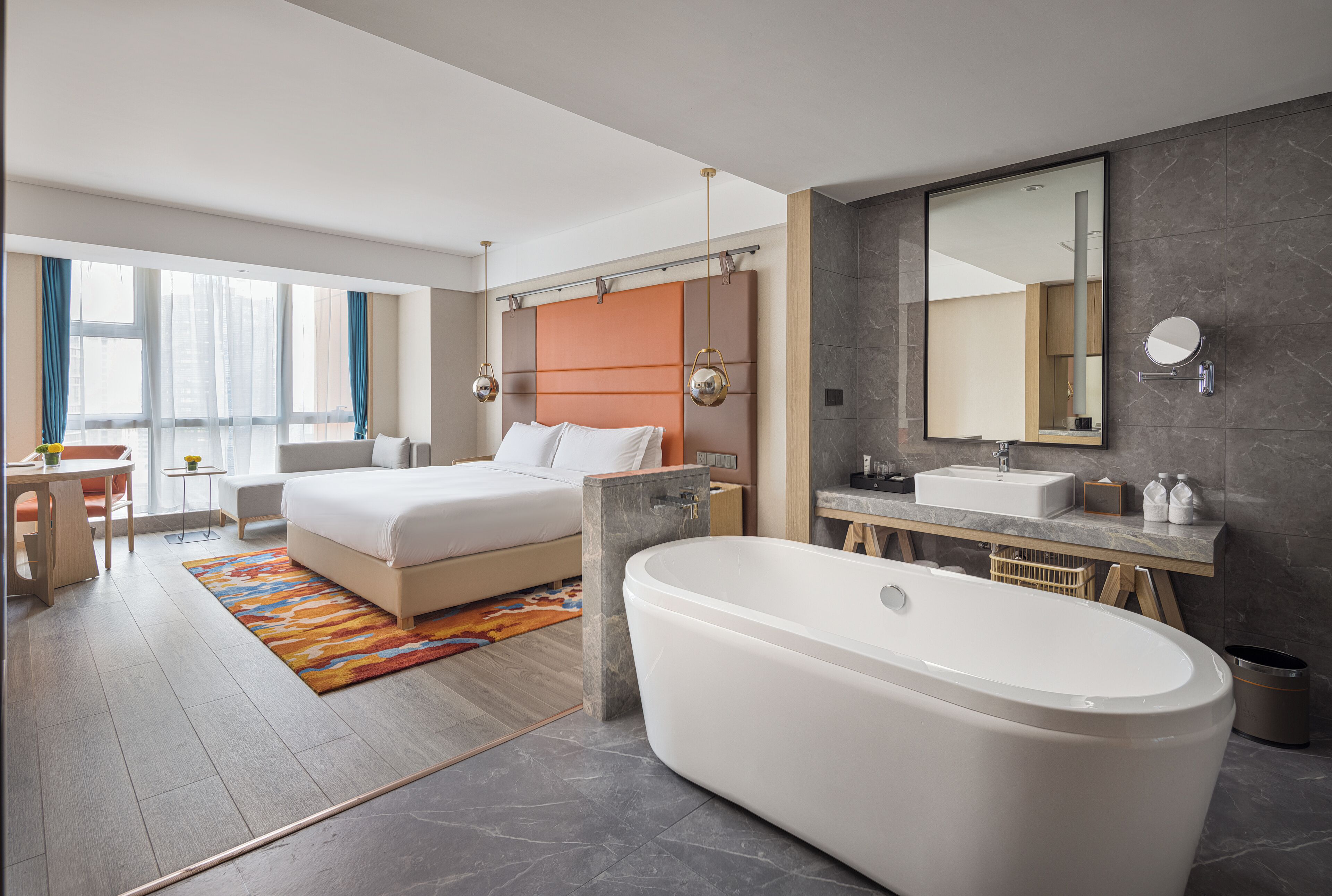 Una habitación de hotel moderna con una cama grande, una zona de estar separada, un baño con bañera y un amplio ventanal que proporciona luz natural.
