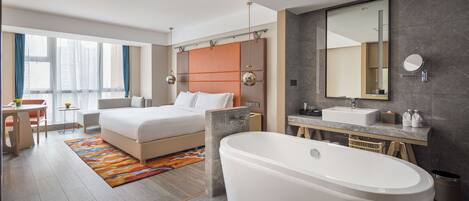Ein modernes Hotelzimmer mit einem großen Bett, einer separaten Sitzecke, einem Badezimmer mit Badewanne und einem großen Fenster für natürliches Licht.