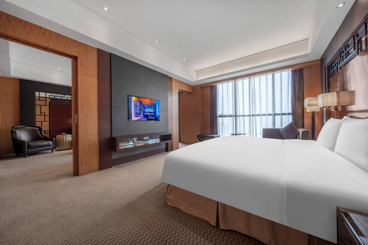 A modern hotel room with a large bed, a flat-screen TV mounted on the wall, a seating area with a chair and a small table, and a large window providing natural light.