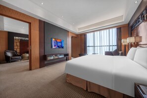 Una habitación de hotel moderna con una cama grande, un televisor de pantalla plana montado en la pared, una zona de estar con una silla y una mesita, y un ventanal que ofrece luz natural.