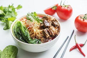 Un tazón de sopa de fideos con carne y vegetales, acompañado de tomates frescos y chiles.