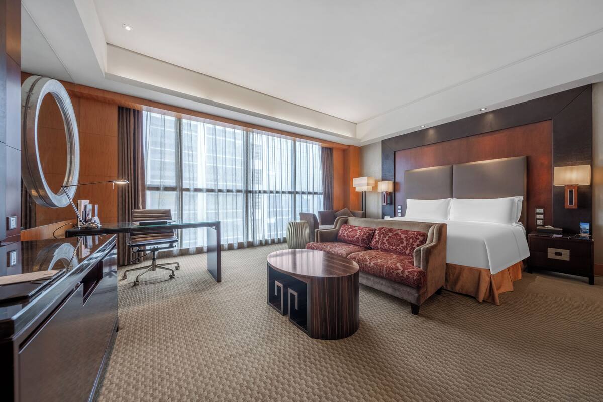 A hotel room with a large bed, a desk, a chair, a round coffee table, and a large window with curtains.