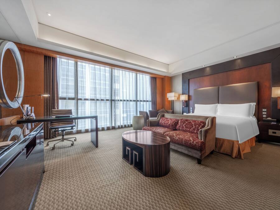 A hotel room with a large bed, a desk, a chair, a round coffee table, and a large window with curtains.