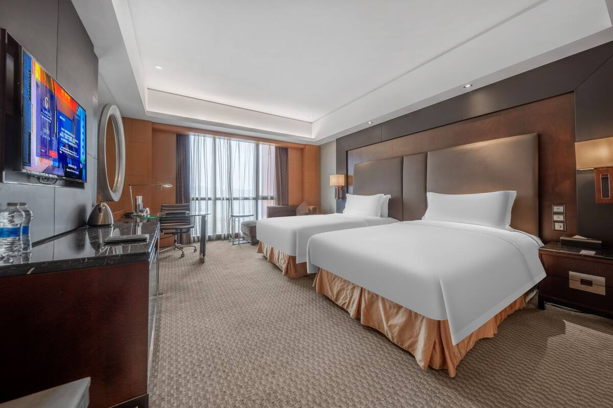 A hotel room with two beds, a desk with a TV, a large window, and a modern design.