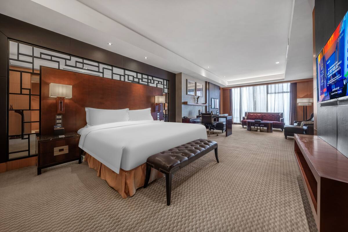 A hotel room with a large bed, a bench, a desk, a TV, and a seating area.