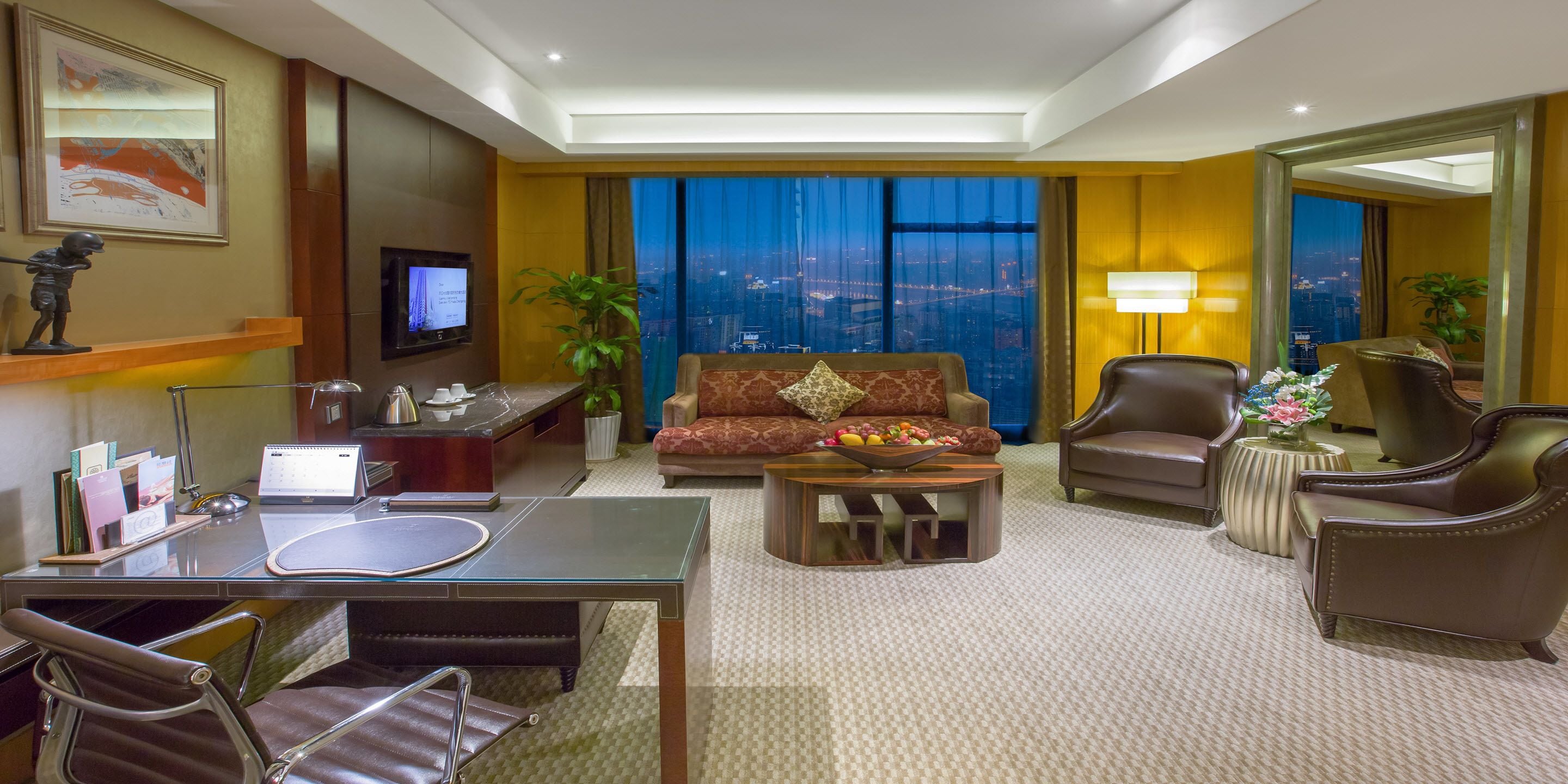 Habitación de hotel con sofá, sillones, mesa de comedor, televisor y vista a la ciudad.