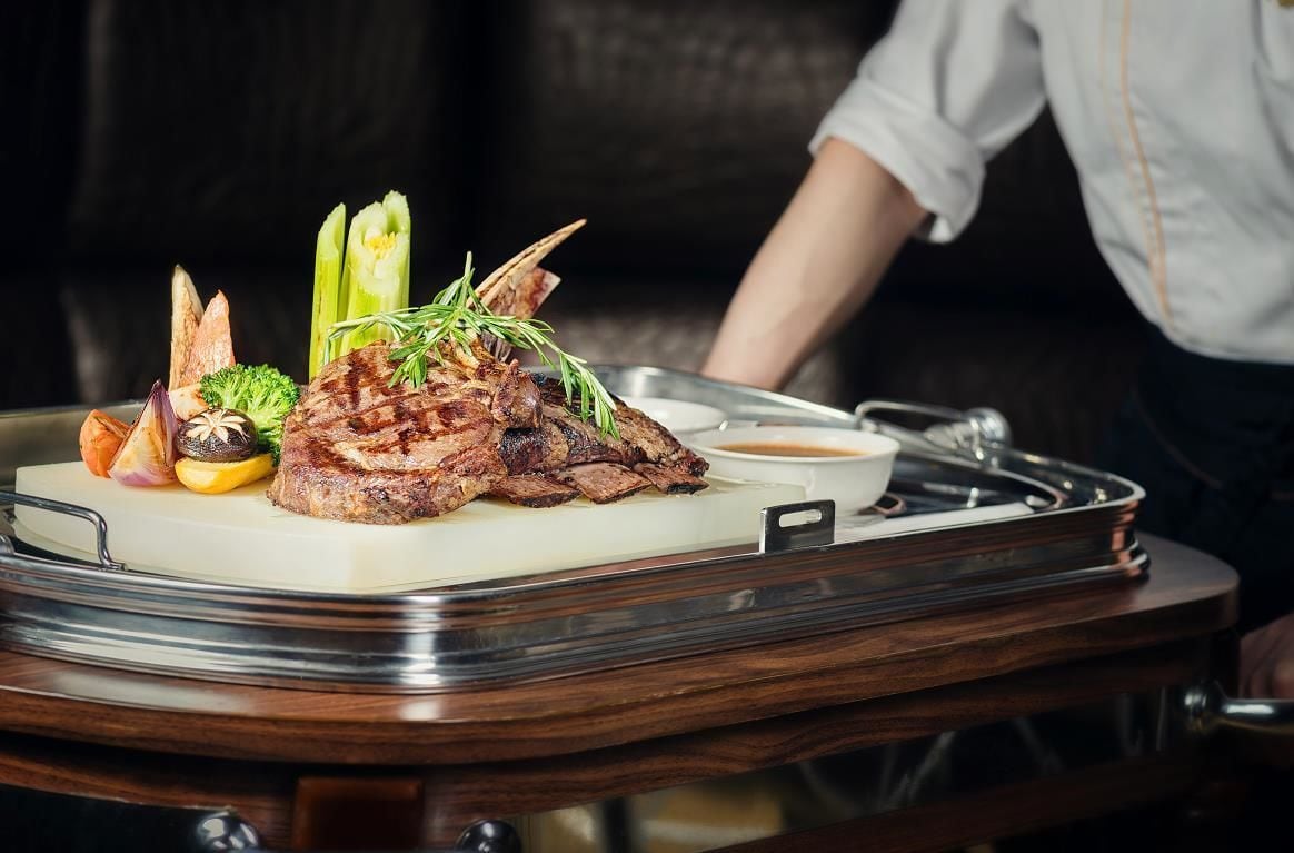 A tray with grilled meat, vegetables, and a small bowl of sauce on a wooden table.
