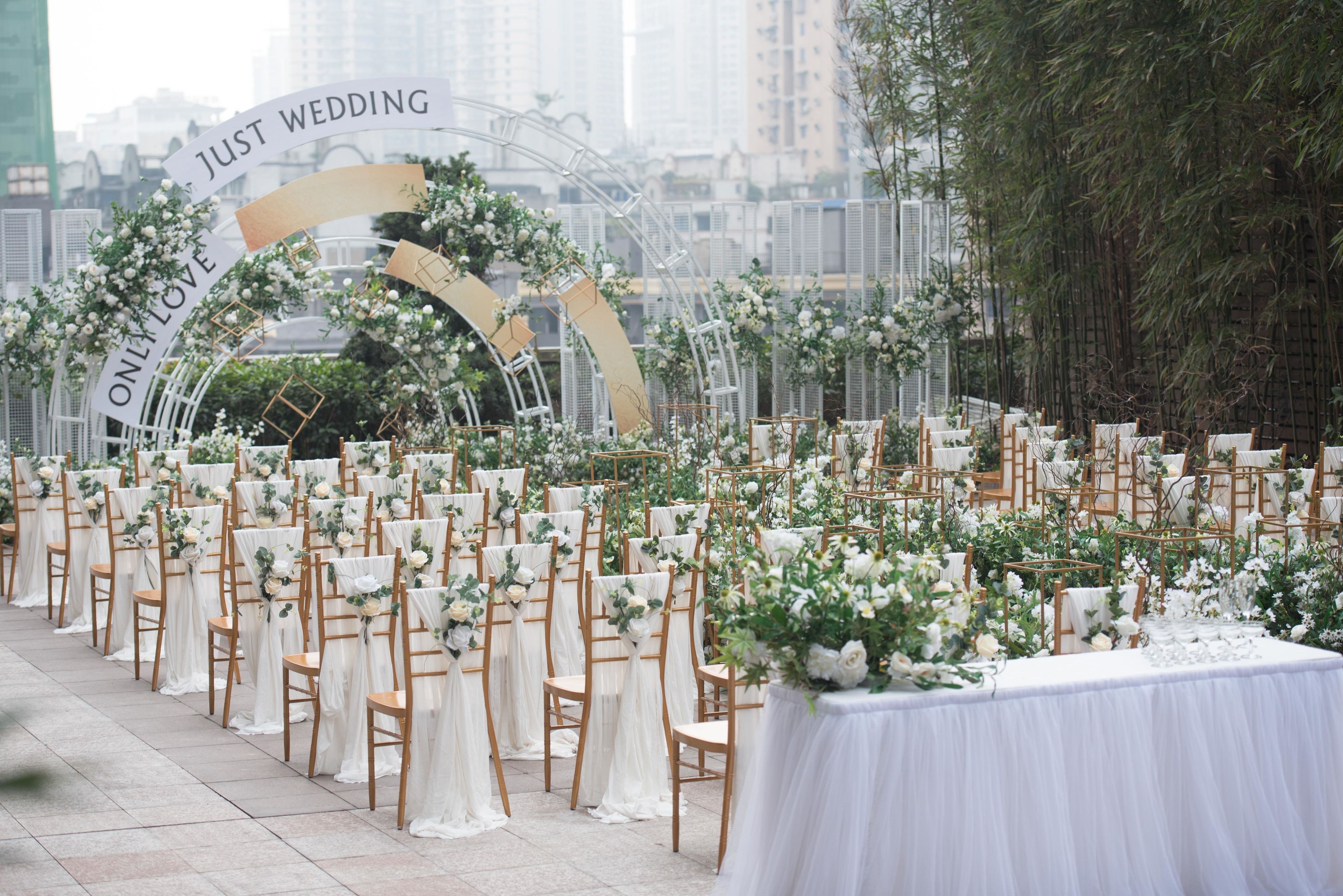 An outdoor wedding venue with rows of wooden chairs and white drapes, a floral arch, and a white tablecloth-covered table.
