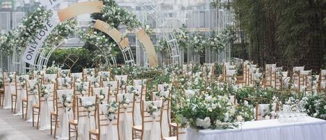 An outdoor wedding venue with rows of wooden chairs and white drapes, a floral arch, and a white tablecloth-covered table.