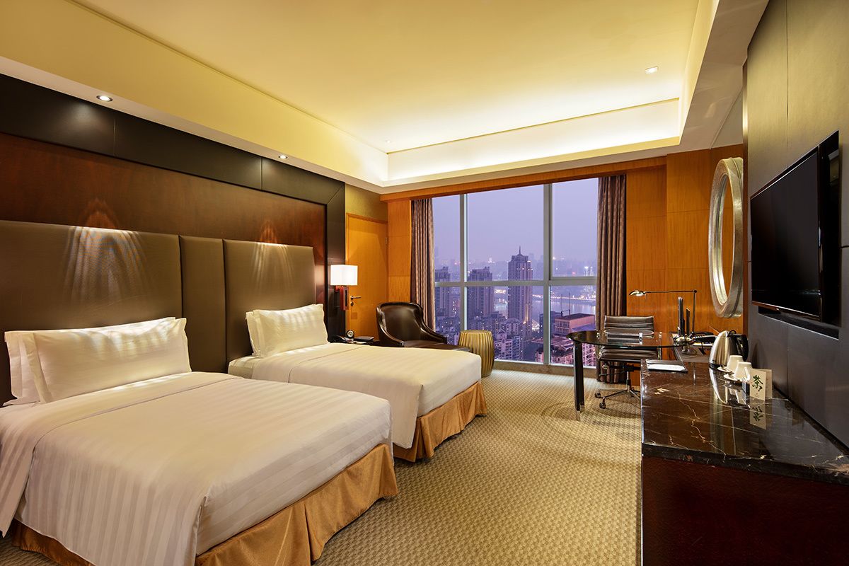 A hotel room with two beds, a desk, a chair, a TV, and a large window with a city view.