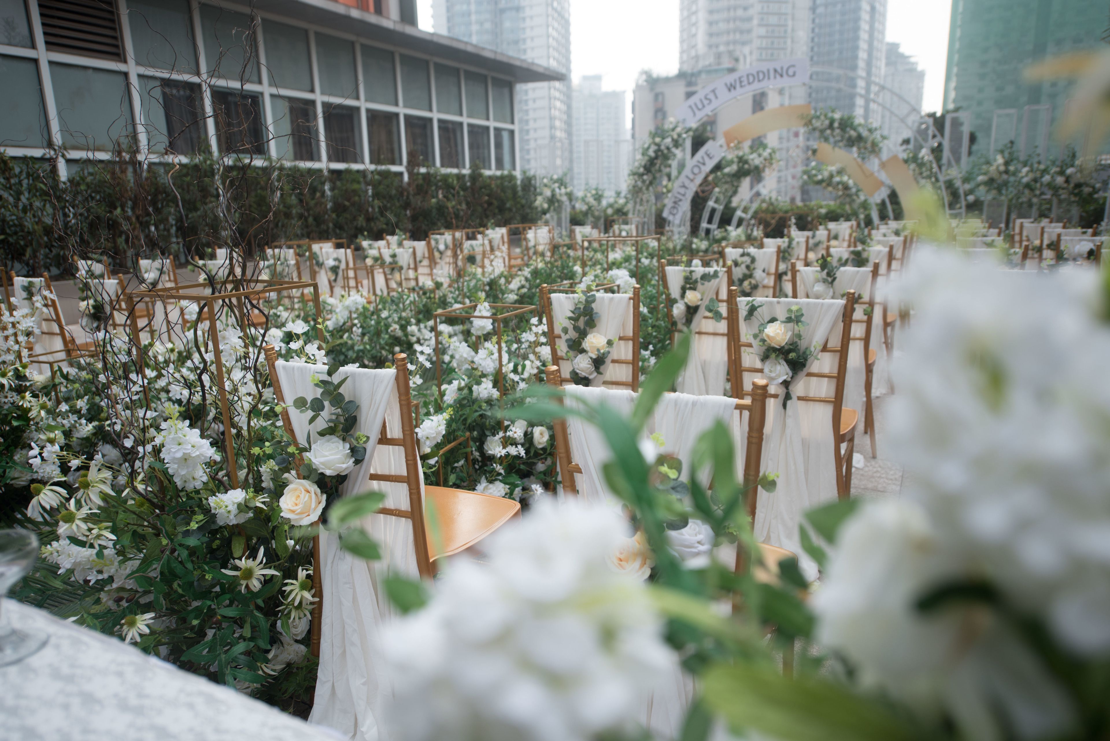Location per matrimonio all'aperto con file di sedie e tavoli di legno, immersa nel verde e circondata da fiori bianchi.