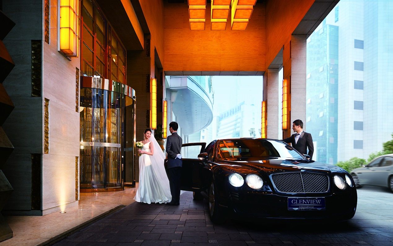 A black Bentley car parked outside a hotel entrance with a glass facade and warm lighting.