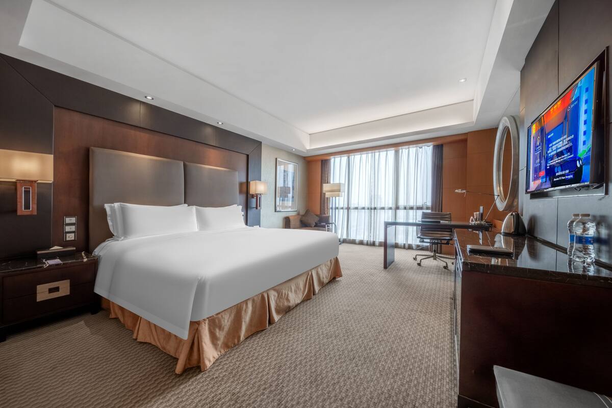 A hotel room with a large bed, a desk, a chair, a TV, and a large window.