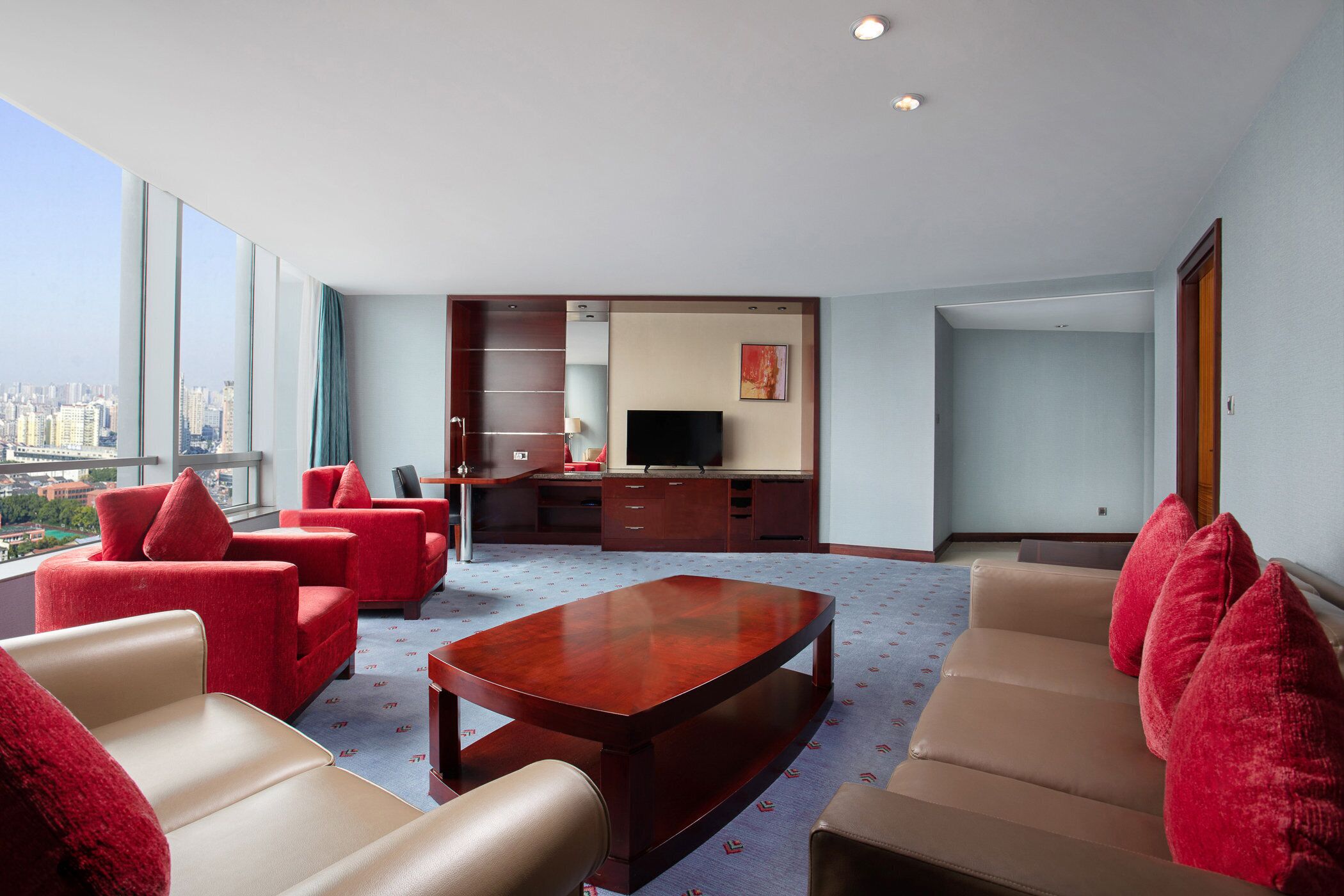 A modern hotel room with a large window, a flat-screen TV, a wooden coffee table, and red and beige sofas.