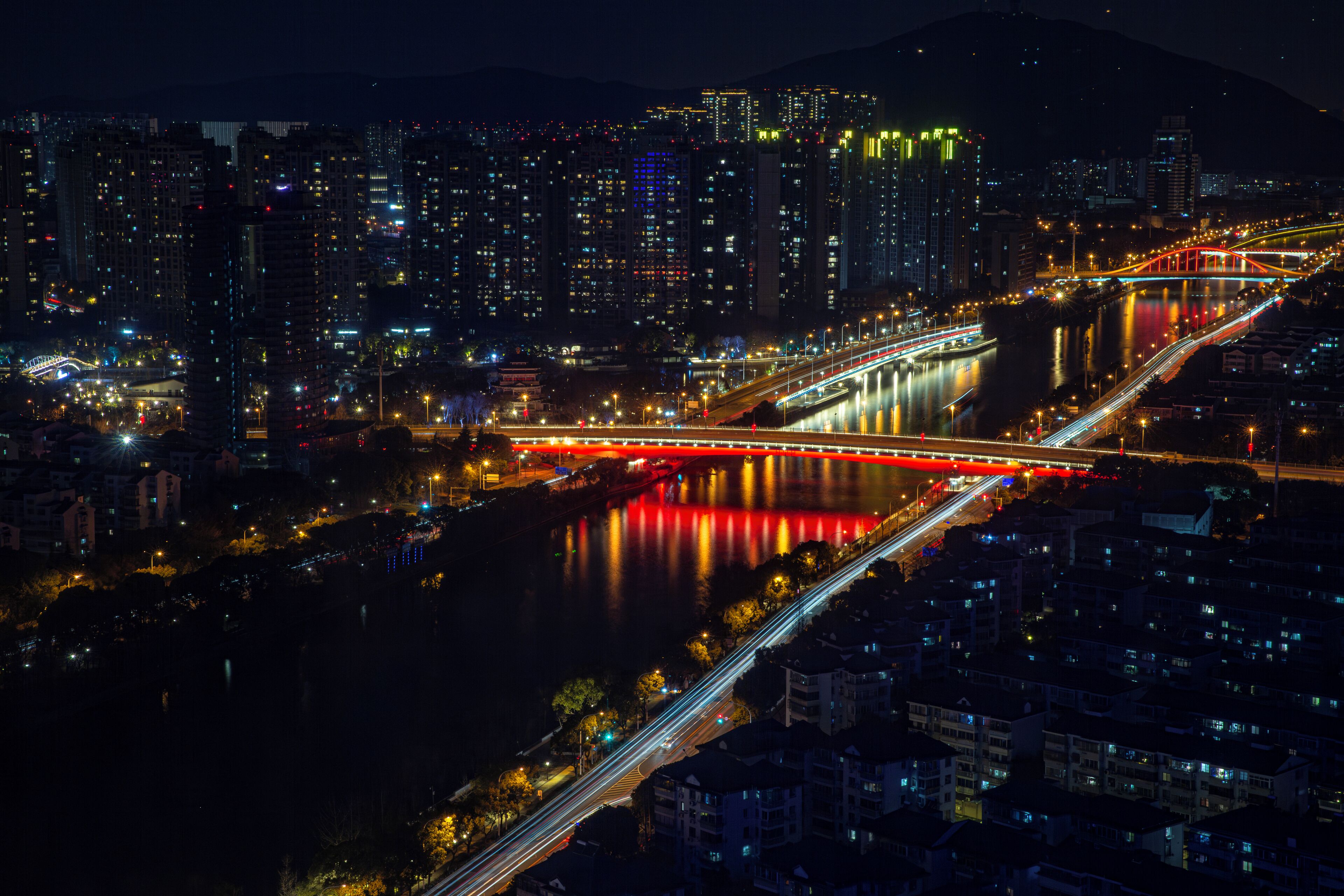 Paisaje urbano nocturno con un puente rojo destacado sobre un río, rodeado de edificios altos y calles iluminadas.