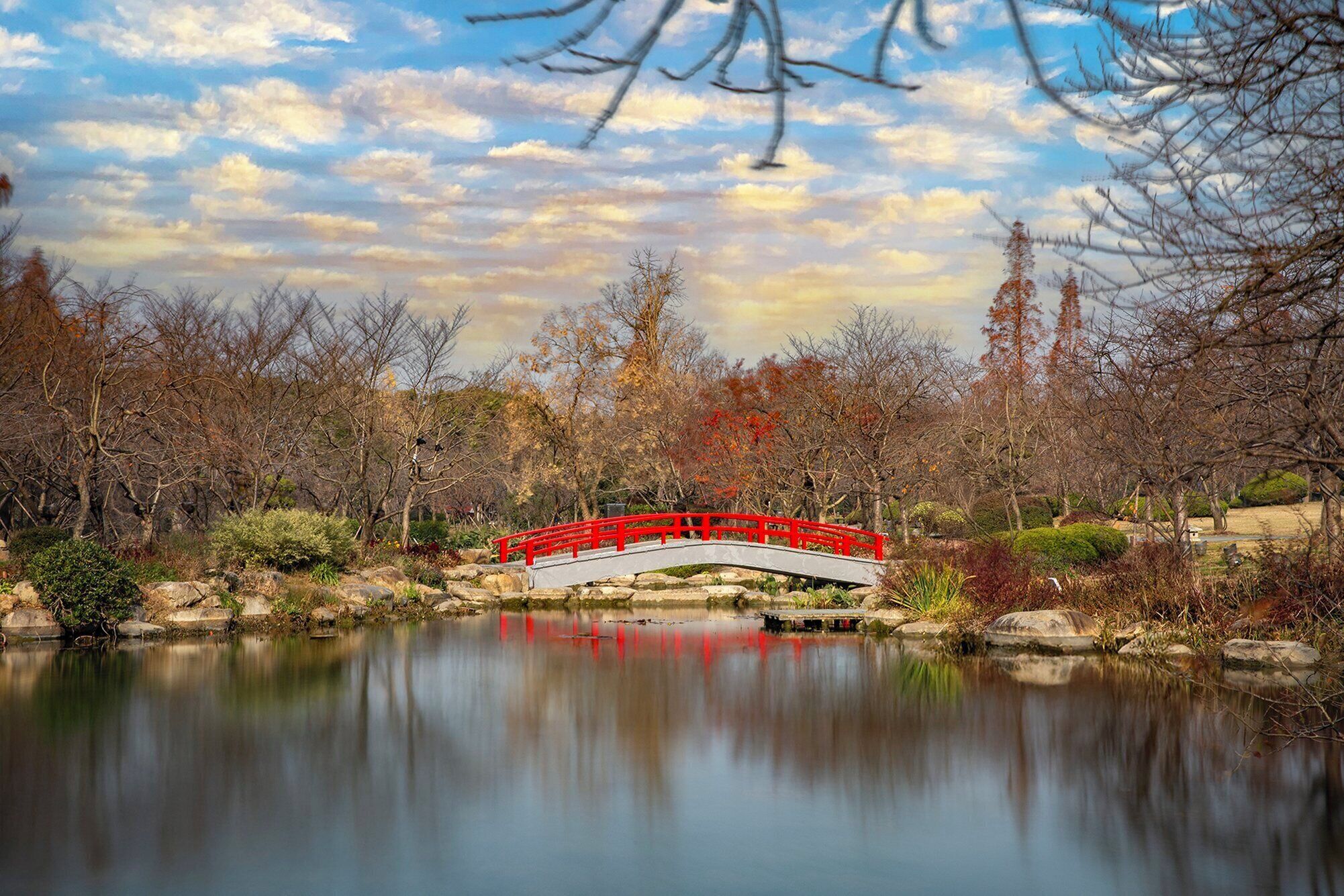 A serene park with a red bridge over a calm lake, surrounded by trees and shrubs.