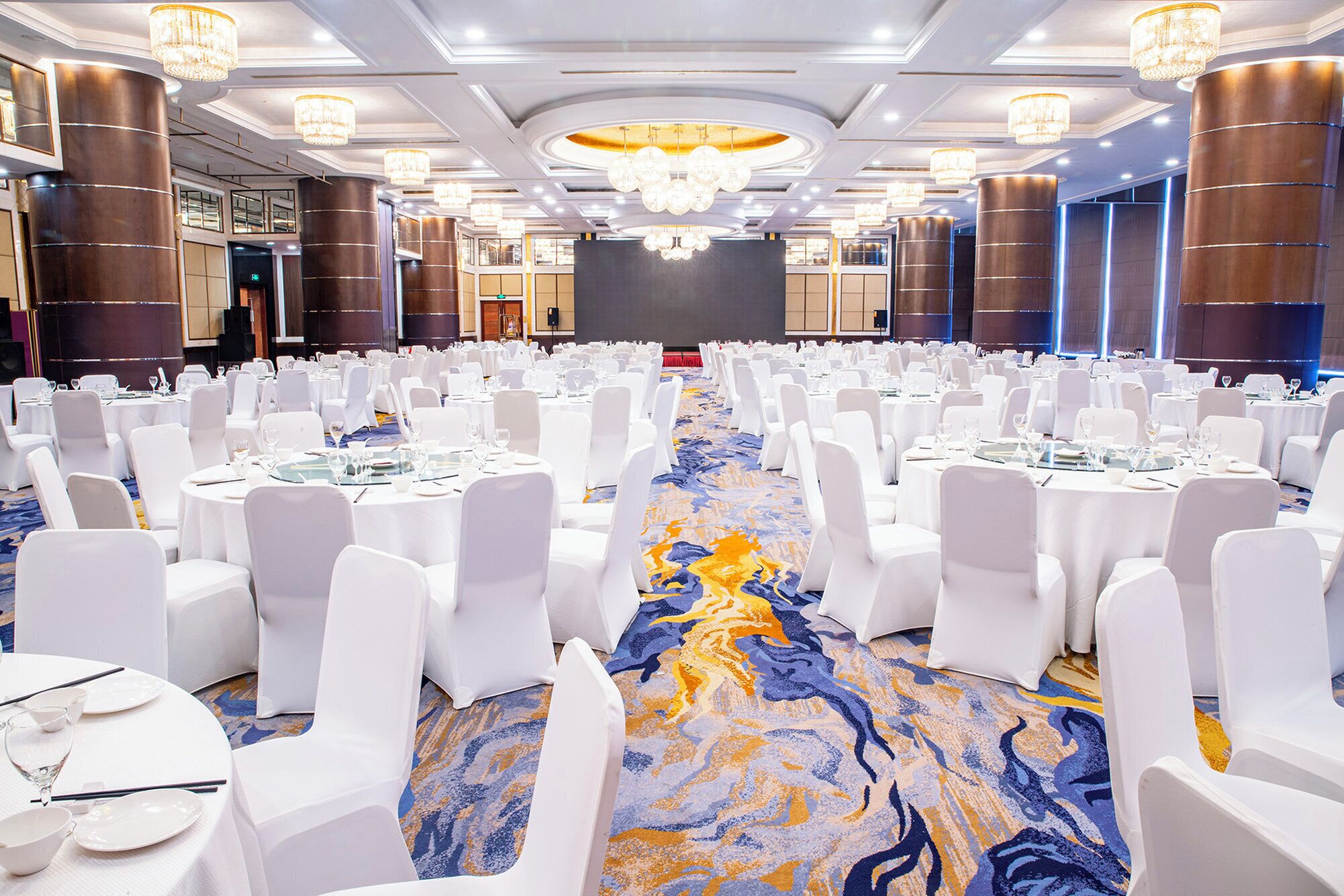 Un salón de banquetes con filas de mesas redondas, sillas blancas y un escenario central.