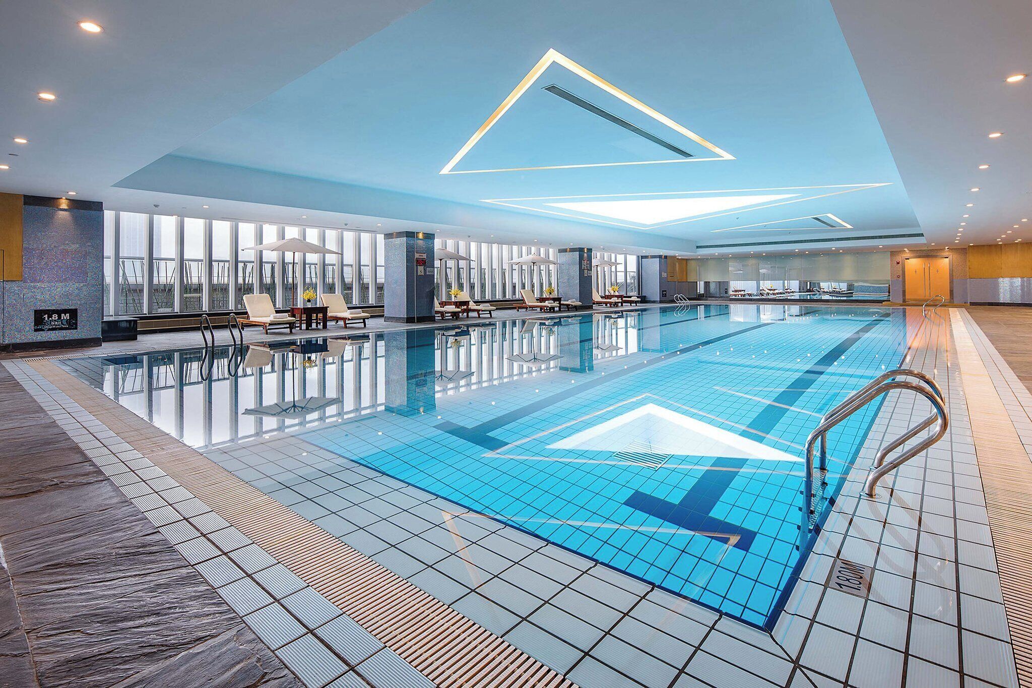 Una amplia piscina interior con agua cristalina, rodeada de sillas de descanso y sombrillas.