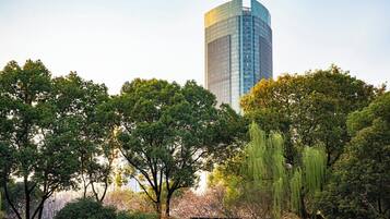 A tall modern building with a glass facade, surrounded by lush green trees and a small structure in the foreground.