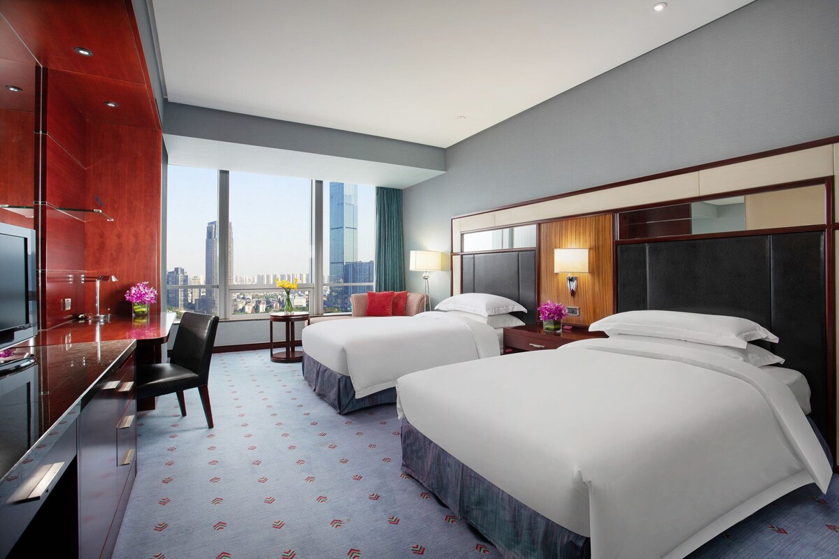 A hotel room with two beds, a desk, a TV, and a large window with a city view.