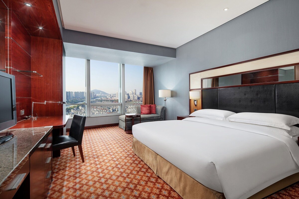 A hotel room with a large bed, a desk, a chair, a TV, and a view of the city.