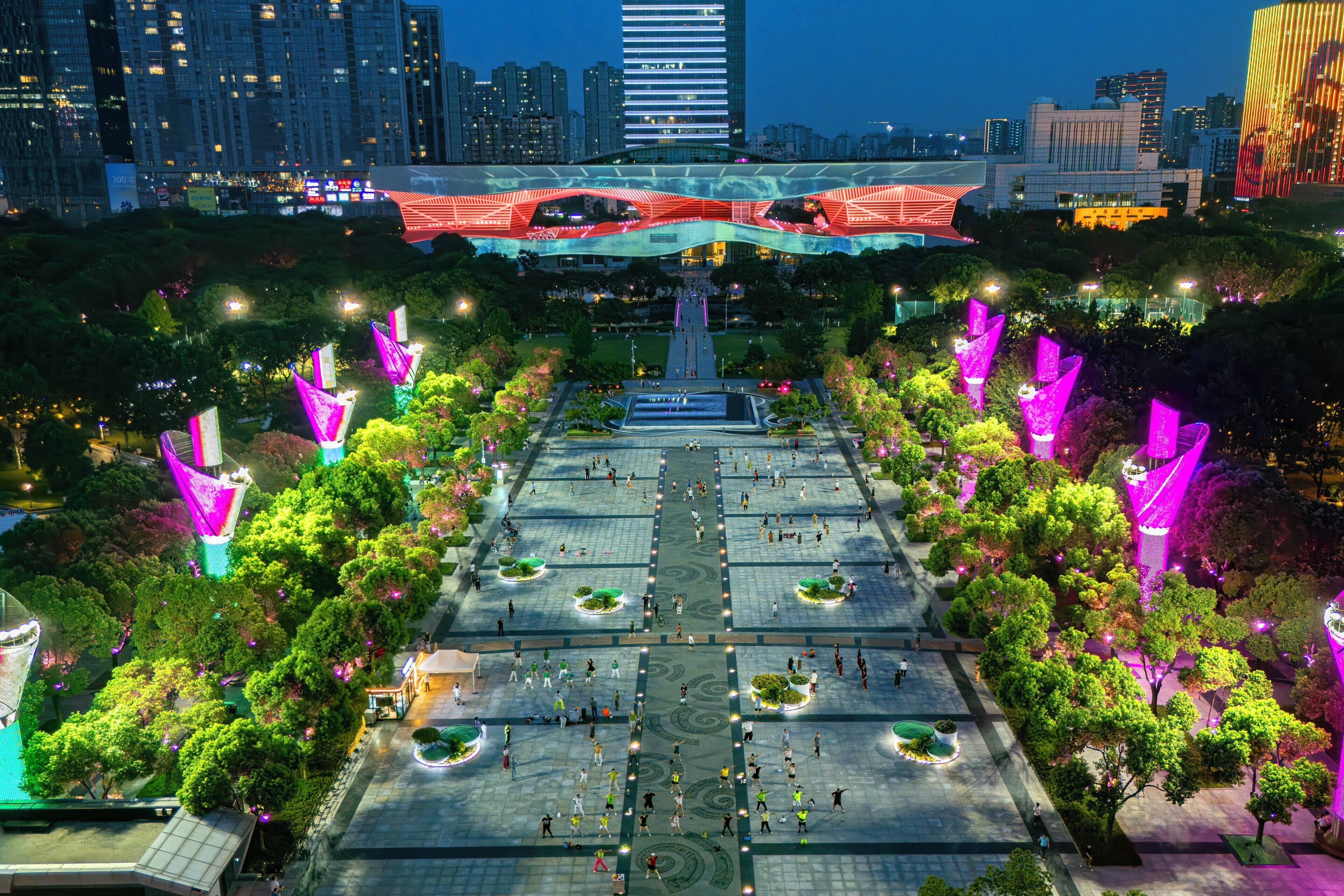 A spacious plaza with a central fountain, surrounded by trees and illuminated by colorful lights.