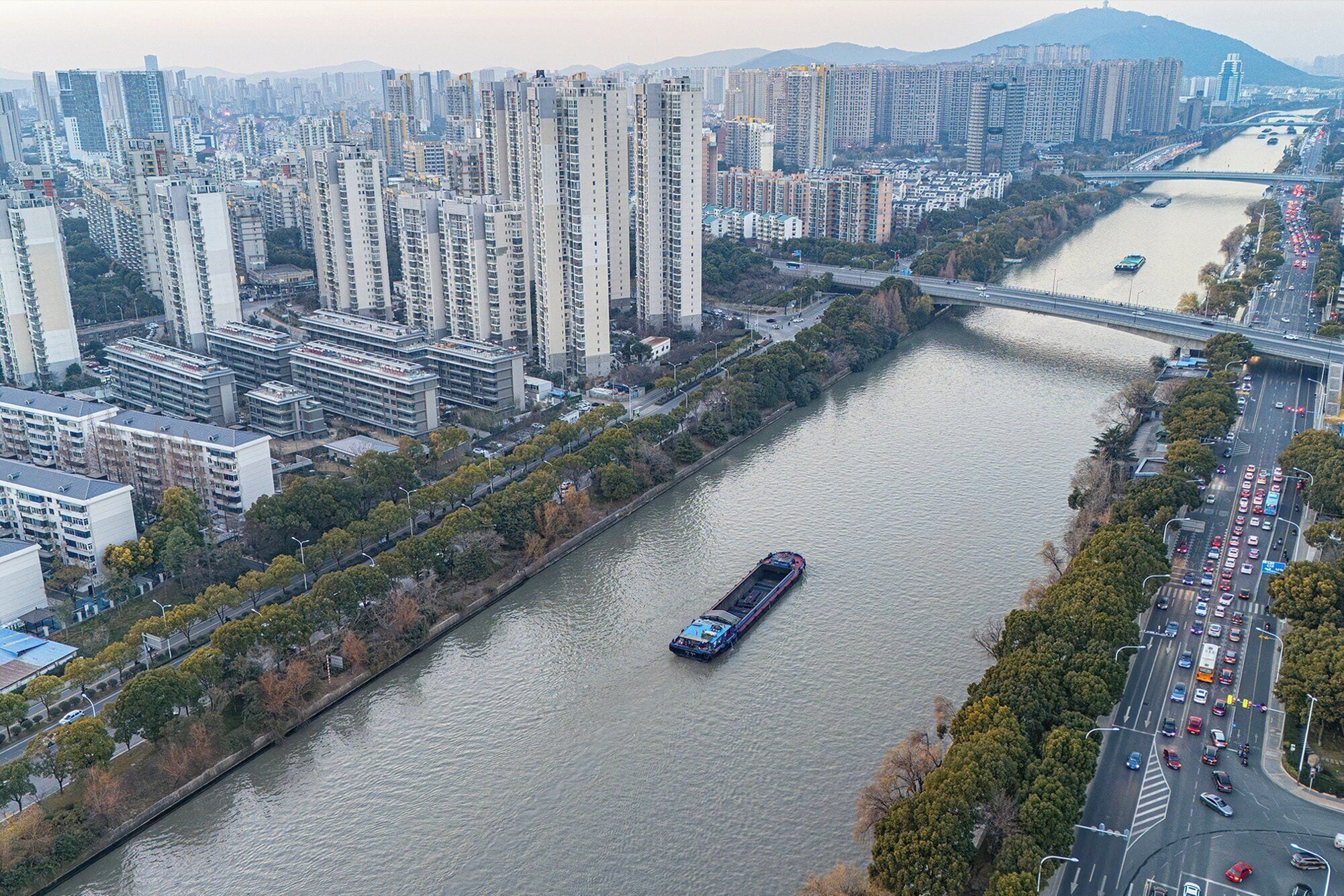 A river with a cargo ship, surrounded by high-rise buildings and a busy road with vehicles.
