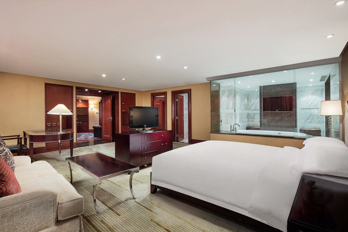 Habitación de hotel con una cama grande, un escritorio, un televisor y vistas de la ciudad a través del ventanal.