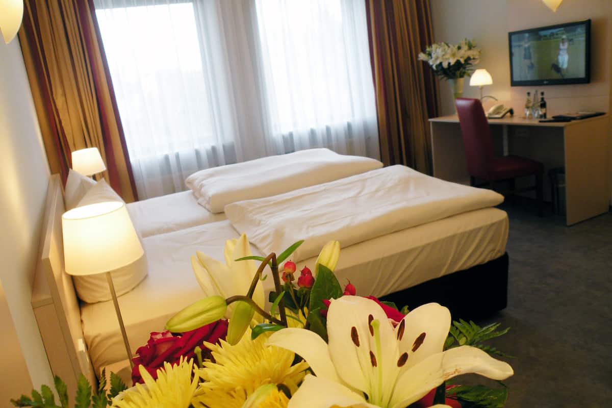 A hotel room with two beds, a desk, a chair, a TV, and a bouquet of flowers.