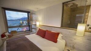 A bedroom with a large bed, wooden headboard, two bedside lamps, a window with a view of a body of water and mountains, and a small table with a vase of flowers. - Noi Indigo Patagonia (Natales)