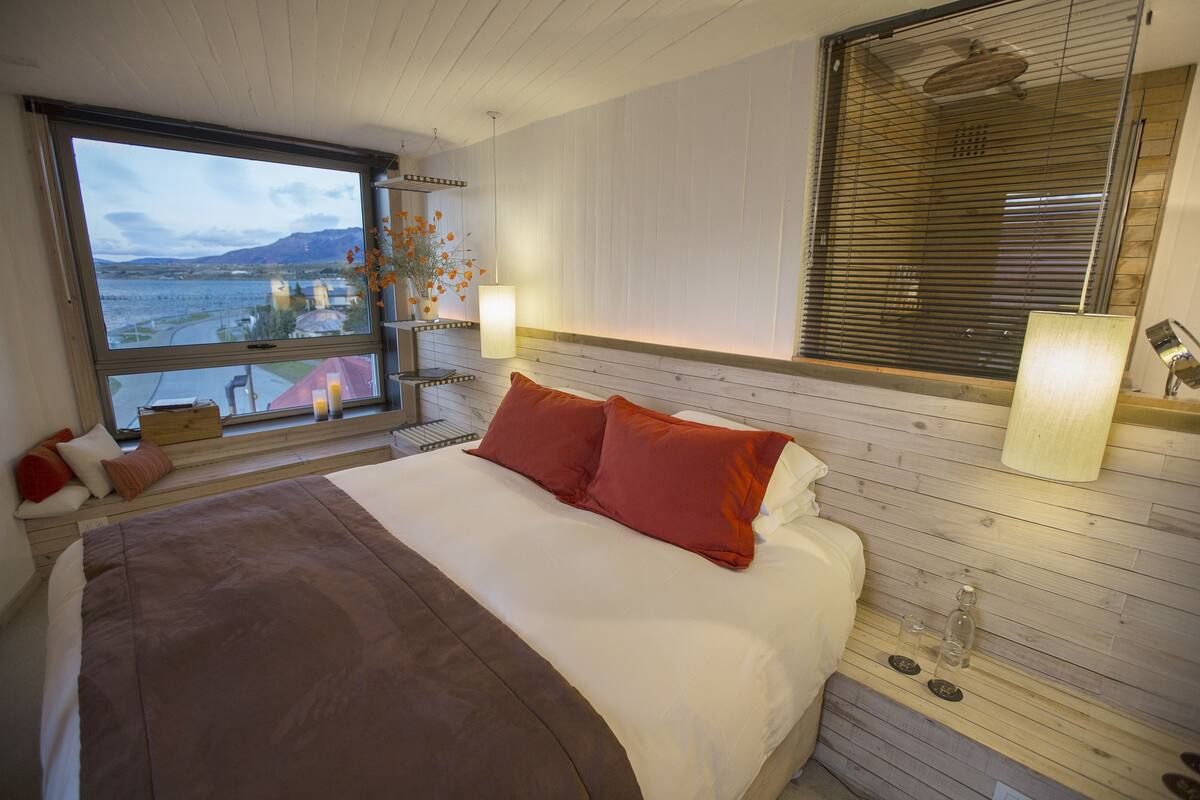 Ein Schlafzimmer mit einem großen Bett, einem hölzernen Kopfteil, zwei Nachttischlampen, einem Fenster mit Blick auf ein Gewässer und Berge sowie einem kleinen Tisch mit einer Blumenvase.