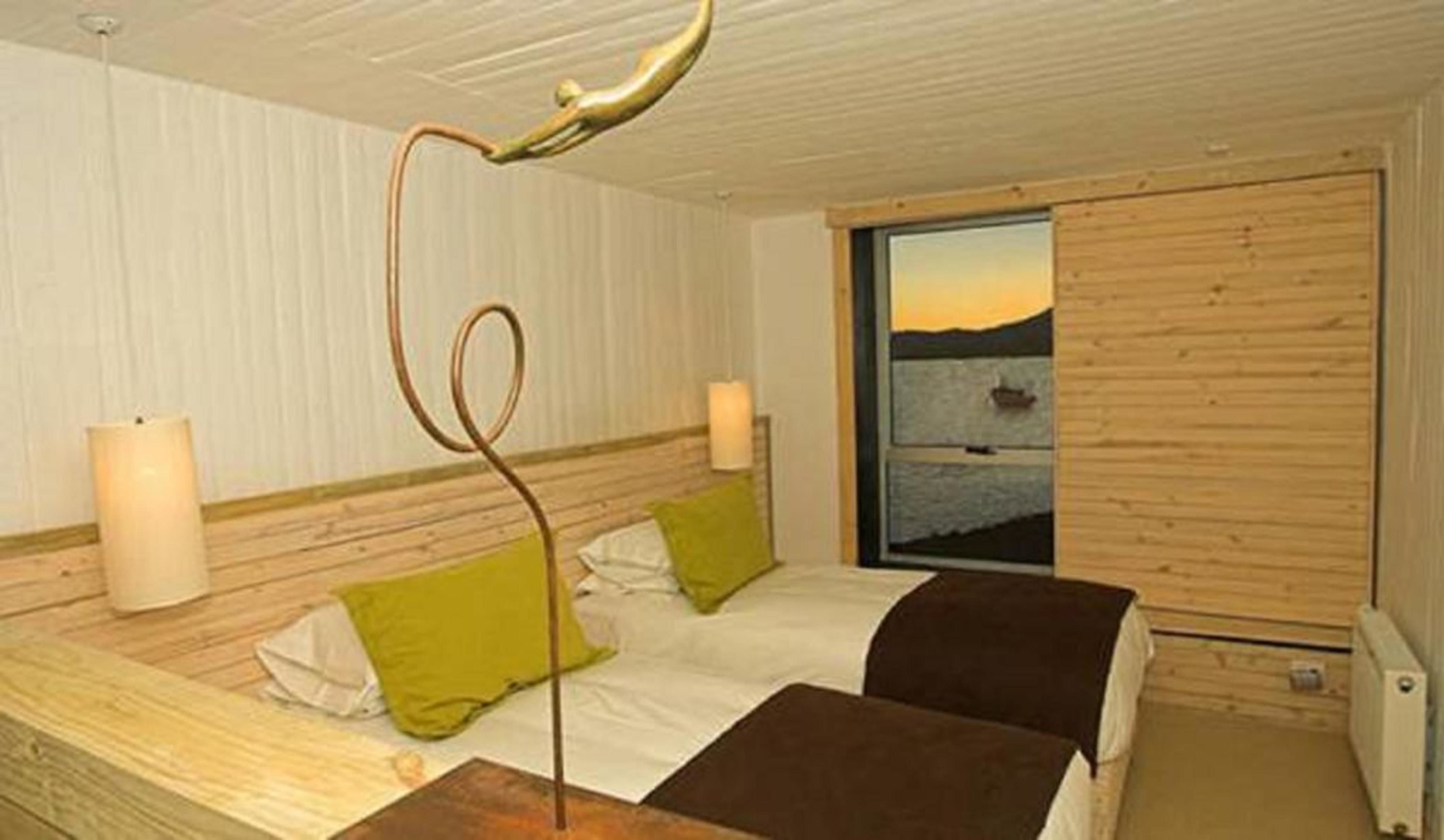Een slaapkamer met een houten hoofdbord, een bed met wit en groen beddengoed, een nachtkastje, een lamp en een raam met uitzicht op een waterpartij.