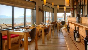 Ein Restaurant mit Holztischen und -stühlen, großen Fenstern mit Blick auf ein Gewässer und hängenden Pendelleuchten.