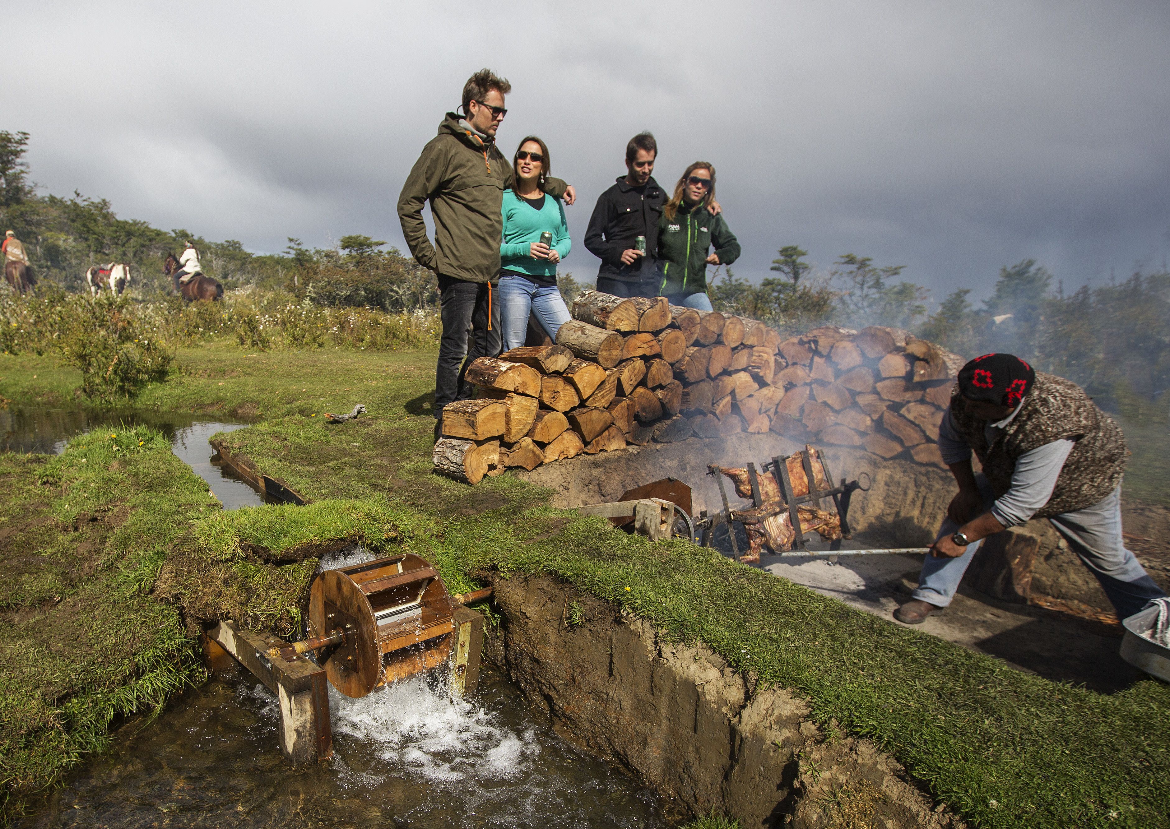 A rural outdoor scene with a water pump, a stack of firewood, and a person grilling meat.