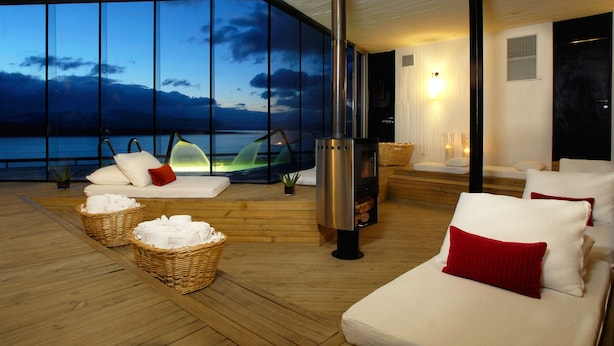 Ein gemütlicher Loungebereich mit einem großen Fenster mit Blick auf ein Gewässer, einem Kamin und bequemen Sitzgelegenheiten.