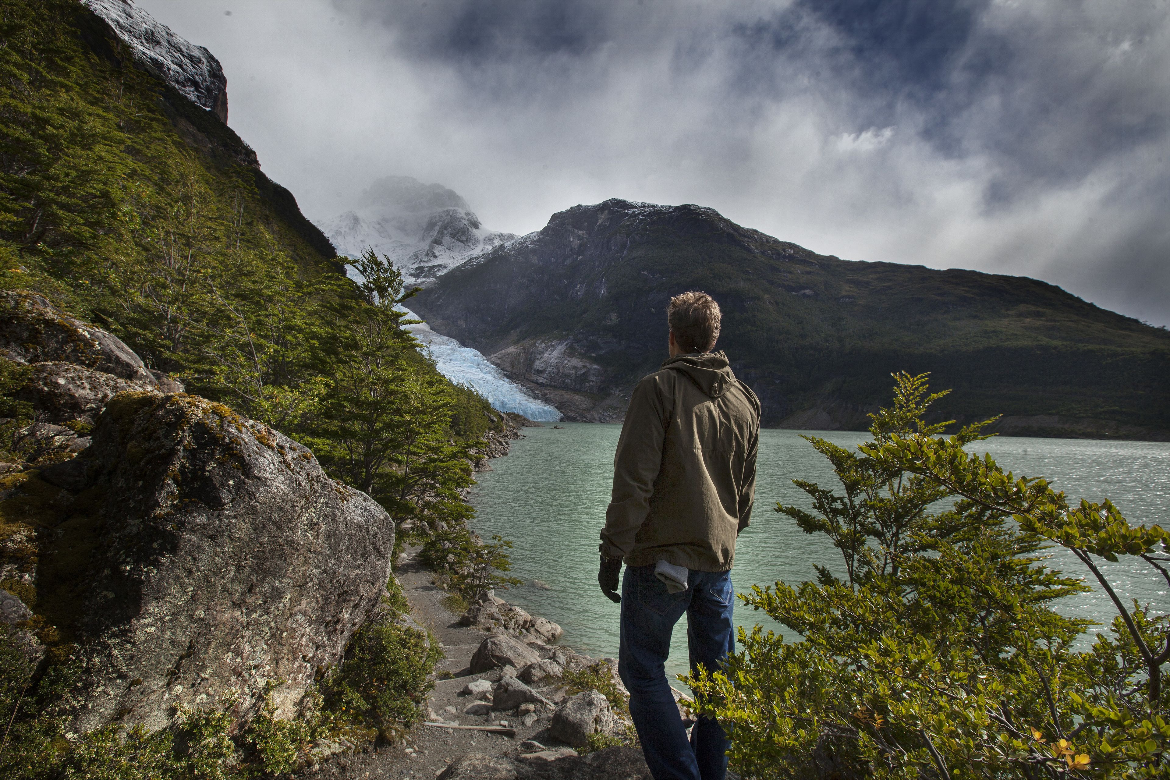 Una persona in piedi su un sentiero roccioso con vista su un ghiacciaio e un lago, con montagne sullo sfondo.
