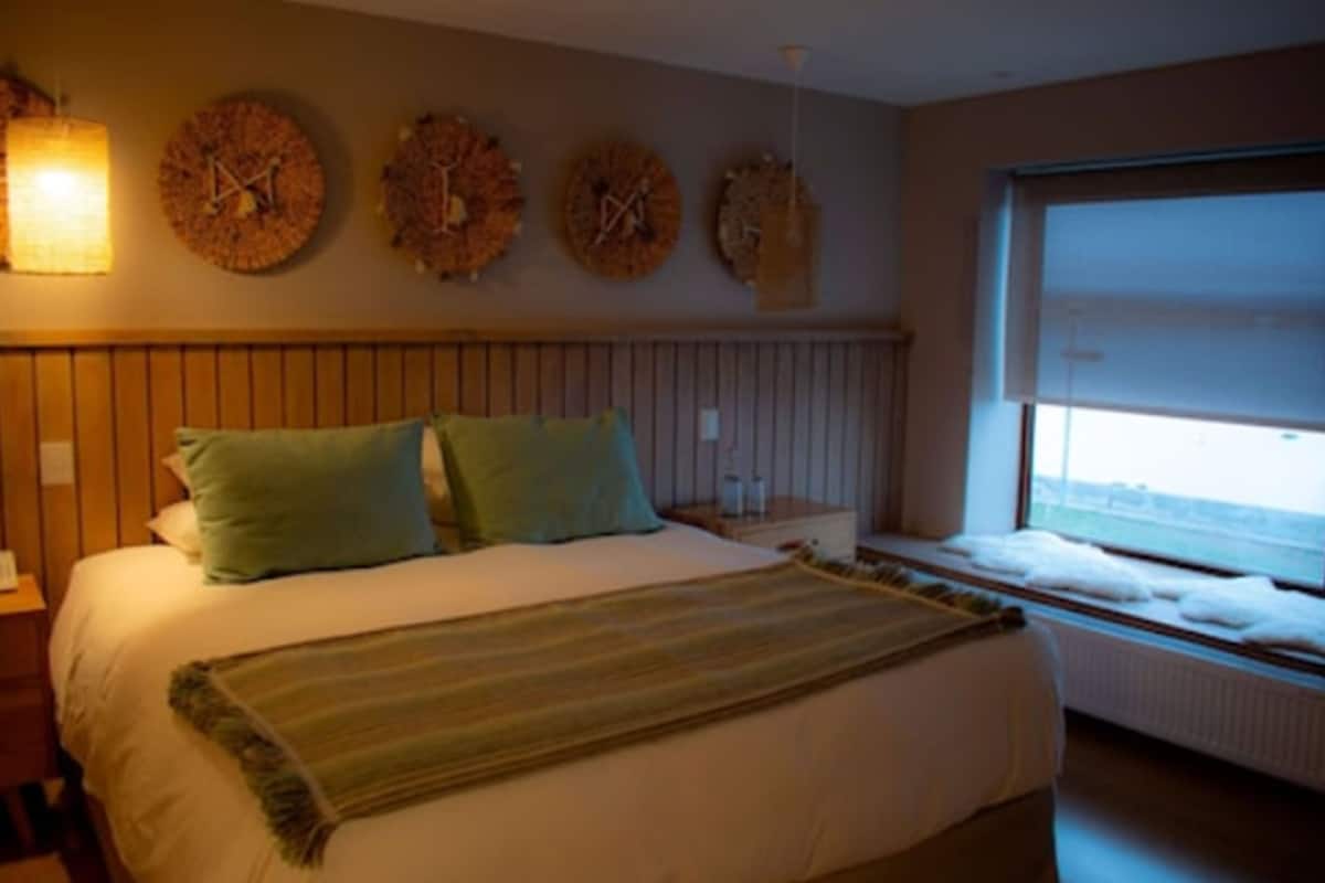 Ein Schlafzimmer mit Bett, Kissen, Decke, einer Wanduhr, einem Fenster und einer Heizung.