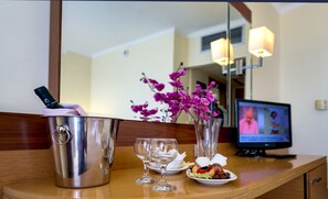 Eine Minibar mit einer Flasche, zwei Weingläsern, einer Obstplatte, einer Vase mit Blumen und einem Fernseher im Hintergrund.