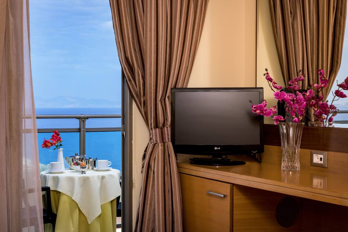 A room with a TV on a wooden cabinet, a table with a white tablecloth, a vase with pink flowers, and a view of the sea through curtains.