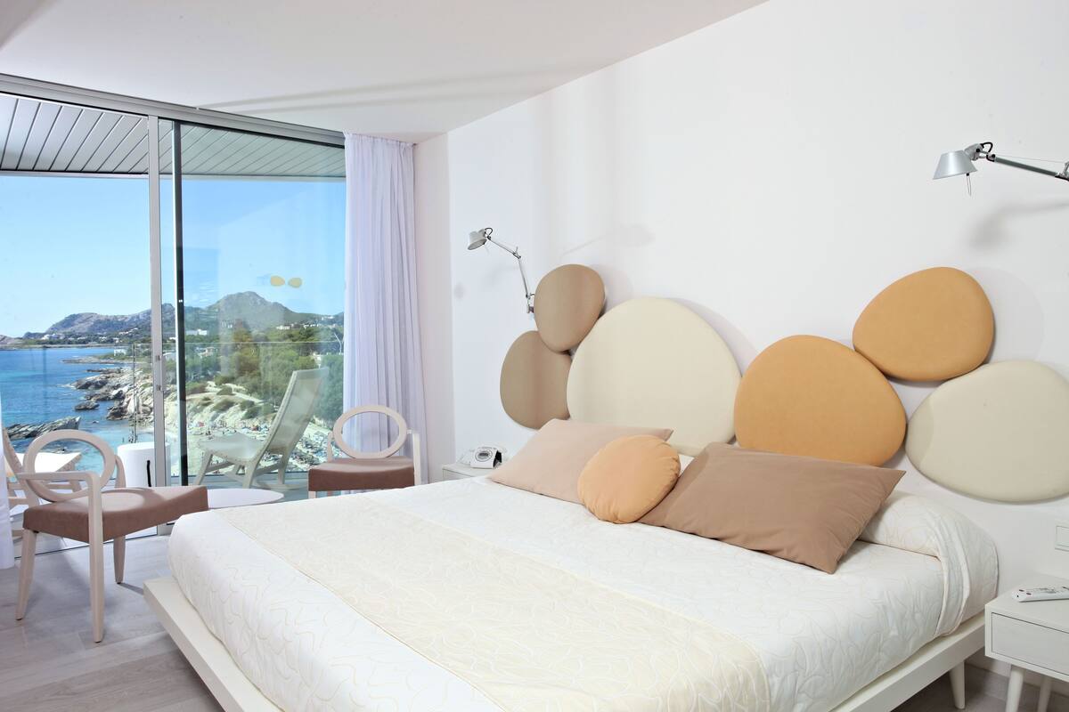 A modern bedroom with a large bed, a glass wall with a view of the sea, and a minimalist design.