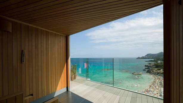 A modern balcony with wooden flooring and a glass railing overlooking a beach with clear turquoise waters and rocky outcrops. - Son Moll Sentits Hotel & Spa (Capdepera)