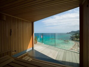 Un balcón moderno con piso de madera y barandilla de vidrio, con vista a una playa de aguas turquesas transparentes y afloramientos rocosos.