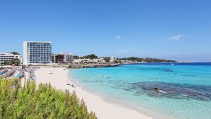 A beach with clear turquoise waters, a sandy shore, and a modern hotel building in the background. - Son Moll Sentits Hotel & Spa (Capdepera)