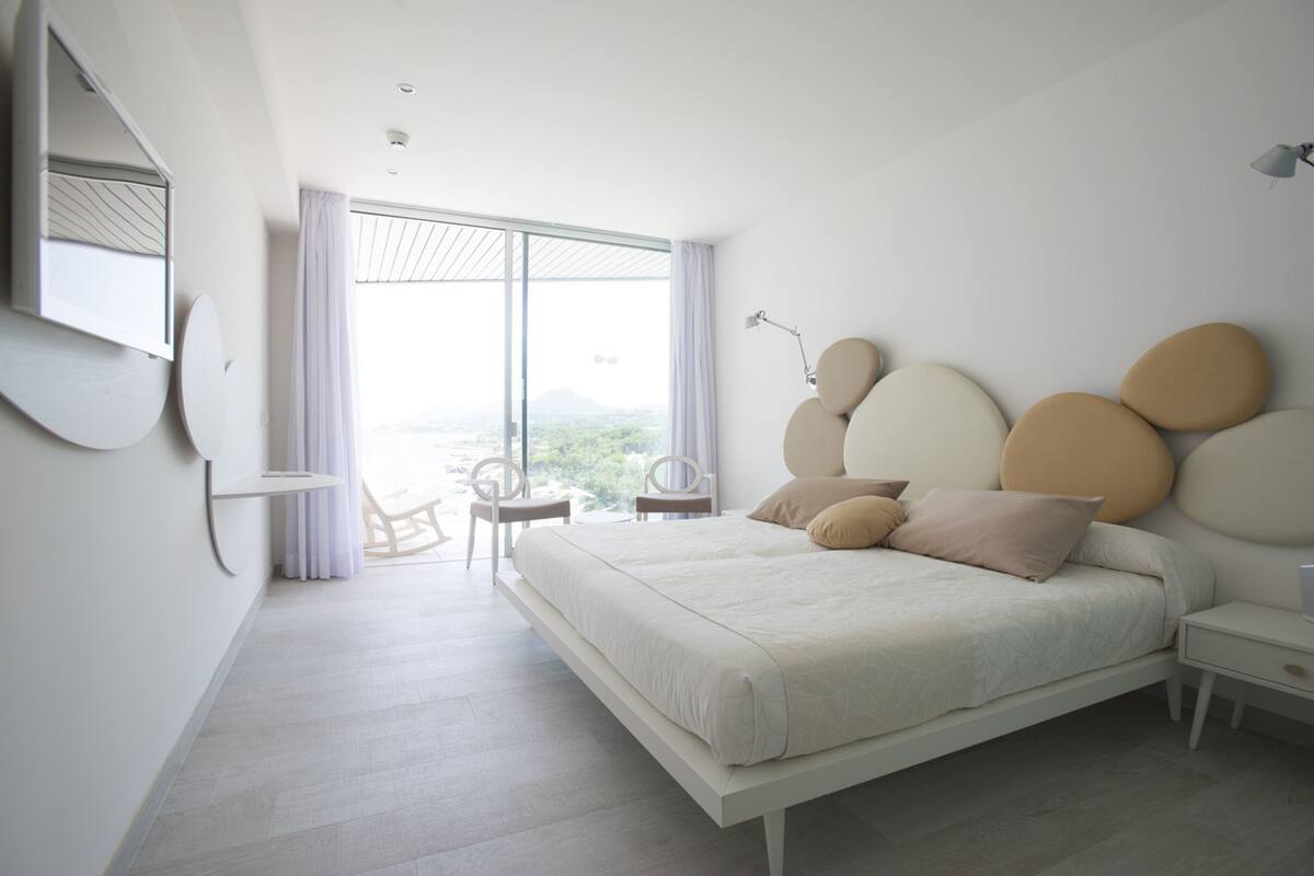 A modern bedroom with a large bed, a bedside table, a chair, and a view of the outdoors.