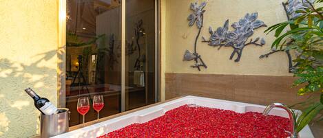 Un baño con una bañera grande llena de pétalos de rosa, una botella de vino y dos copas de vino tinto.