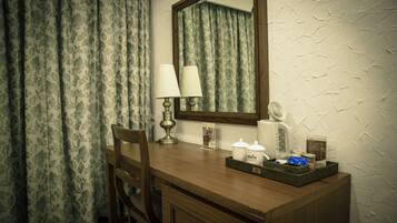 A hotel room with a wooden desk, two chairs, a mirror, a lamp, a tray with toiletries, and a wall with a textured pattern.